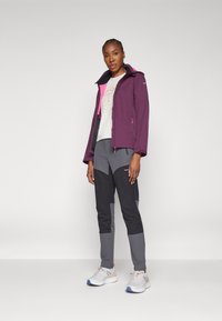 Giacca impermeabile viola con accenti rosa, abbinata a pantaloni grigi e neri a gamba stretta. Scarpe bianche con dettagli arancioni e blu.