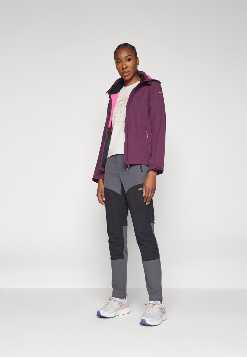 Lila wasserdichte Jacke mit pinken Akzenten, kombiniert mit grauen und schwarzen schmalen Hosen. Weiße Schuhe mit orangefarbenen und blauen Details.