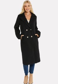 Cappotto doppiopetto nero con grandi bottoni, colletto ampio, tasche laterali e maniche lunghe, abbinato a jeans blu e stivaletti beige.