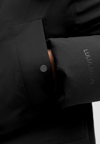 Osoba nosząca czarną kurtkę Lululemon z fakturowanej tkaniny i napem, mankiet rękawa z widoczną nazwą marki.