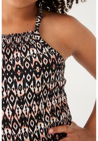 Zbliżenie na osobę ubraną w bezrękawnikową sukienkę o marszczonym fasonie w czarno-brzoskwiniowy geometryczny wzór, ręka spoczywa na biodrze.