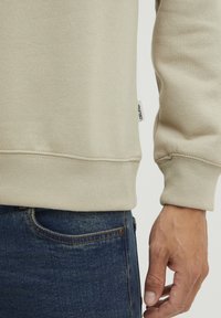 Sudadera beige con puños y dobladillo acanalados, con una pequeña etiqueta de tela. Mano apoyada sobre unos pantalones vaqueros de denim azul oscuro.