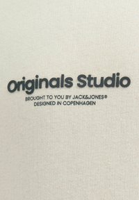 Tessuto beige chiaro con una texture liscia che presenta testo stampato in nero: "Originals Studio", "portato a te da JACK&JONES" e "progettato a Copenaghen."