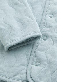 Helles blauer, gestepptes Gewebe mit wellenförmiger Textur; verfügt über einen runden Druckknopf; aus weichem Baumwollmaterial mit gestickten Details entlang der Kanten.