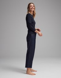 Navy long-sleeve shirt paired with dark denim straight-leg jeans featuring a raw hem. Model stands barefoot, smiling against a neutral gray backdrop.