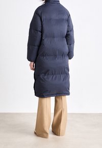 Dunkelblauer Steppmantel mit langer Silhouette, ausgestattet mit einem hohen Kragen, verlängerten Ärmeln und horizontalen Steppnähten.