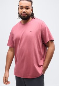 Άνδρας με ντραντλόκ και γενειάδα φορώντας ένα μπλουζάκι Levi’s σε χρώμα ροδακινί και σκούρο παντελόνι, στέκεται μπροστά σε ένα απλό ανοιχτόχρωμο φόντο.