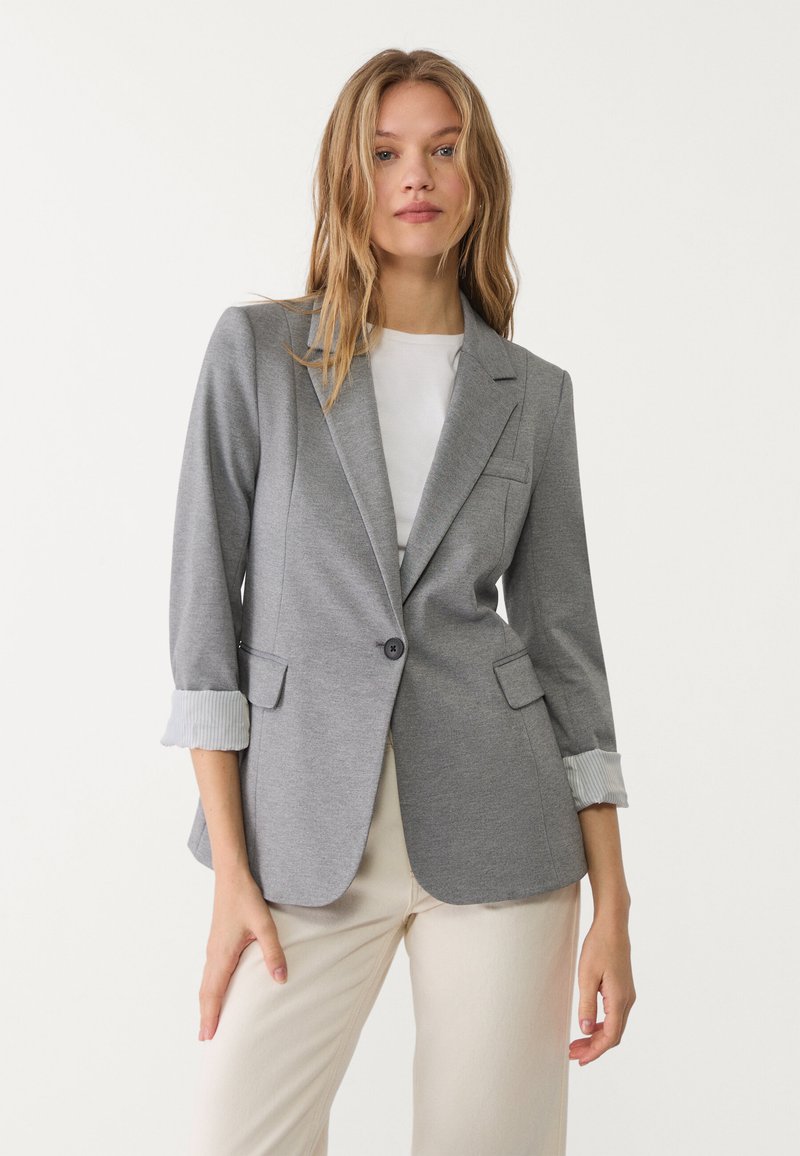 Šedý blazer s přiléhavým střihem, zapínání na jeden knoflík, dvě přední kapsy a ohrnuté rukávy s pruhovaným detailem.