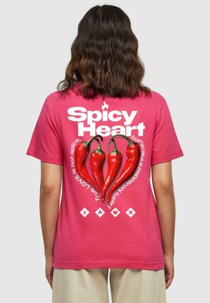Persoon die een roze shirt draagt met rode chilipepers in een hartvorm gerangschikt en de tekst "Pittig Hart" en gerelateerde uitdrukkingen op de achterkant.