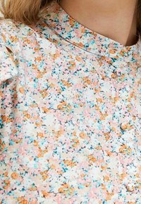 Blusa floreale con sfondo crema chiaro, con motivi floreali rosa, arancioni e blu sparsi, dotata di chiusura a bottoni e colletto alla mandarino.