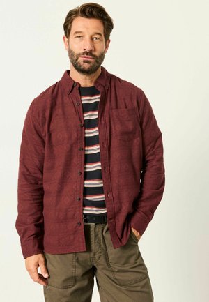 Mann mit Bart, der ein burgunderfarbenes Hemd mit Knöpfen über einem schwarz-weiß-roten, gestreiften T-Shirt und olivgrünen Hosen trägt, steht mit einer Hand in der Hosentasche.