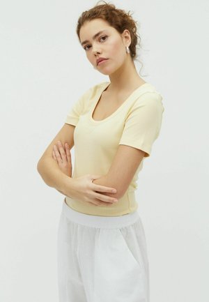 Tânără cu păr creț, purtând un top pal galben cu mâneci scurte și pantaloni albi, stând cu brațele încrucișate pe un fundal simplu.