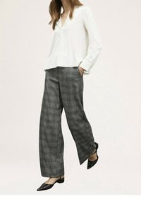 Kobieta w białej bluzce z długim rękawem, szarych szerokich spodniach w kratę i czarnych szpilkach na odkrytej pięcie, idąca z jedną ręką w kieszeni.
