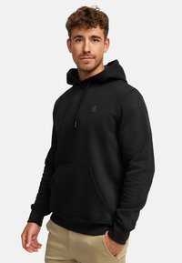 Ung mand med krøllet hår iført en sort hættetrøje og beige bukser, stående med den ene hånd i lommen, neutral baggrund.