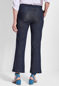Pantalon en denim bleu foncé avec une coupe large. Il dispose d'une taille élastique et de deux poches arrière. Porté avec des sandales à talons hauts bleues.