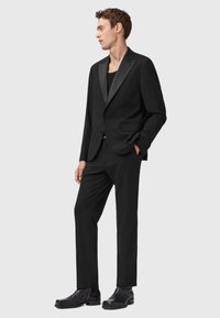 Uomo in piedi che indossa un abito nero con rever in satin, camicia nera, pantaloni eleganti neri e stivaletti in pelle nera, con una mano in tasca.