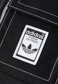 Tessuto nero con un'etichetta cucita bianca con il testo "adidas" e un logo trefoil. L'etichetta ha una forma rettangolare con angoli arrotondati.
