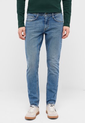 Mann trägt grünes Langarmhemd, blaue Slim-Fit-Jeans und weiße Sneakers, steht vor einfachem hellem Hintergrund.