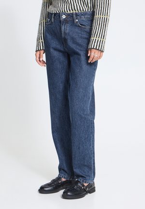 Personne portant un jean droit bleu foncé, des mocassins en cuir tressé noirs, un haut rayé à manches longues et des bagues argentées aux doigts.