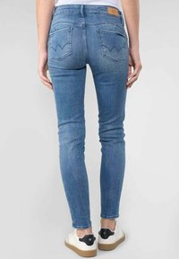 Vista trasera de una persona que lleva jeans ajustados de mezclilla azul claro y zapatillas blancas con detalle negro en el talón, de pie sobre un fondo liso.