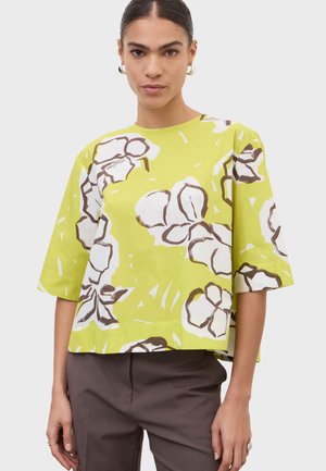 Kobieta ubrana w luźną limonkowo-zieloną bluzkę z dużymi białymi i brązowymi kwiatowymi nadrukami oraz rękawami do łokcia, w połączeniu z ciemnobrązowymi spodniami.