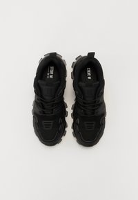 Chaussures de sport noires avec une tige en mesh texturé, un col rembourré et une semelle épaisse à crampons. Présente un logo en relief sur la languette et sur le côté.