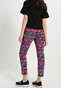 Černý top s krátkými rýhami na rukávech, spárovaný s kalhotami s mnohobarevným geometrickým vzorem v růžové, zelené, oranžové a černé. Černá obuv.