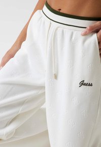 Białe ribbed jogger pants z elastycznym zielonym pasem, haftowanym logotypem w brązie oraz subtelnymi detalami teksturowymi na całej powierzchni.