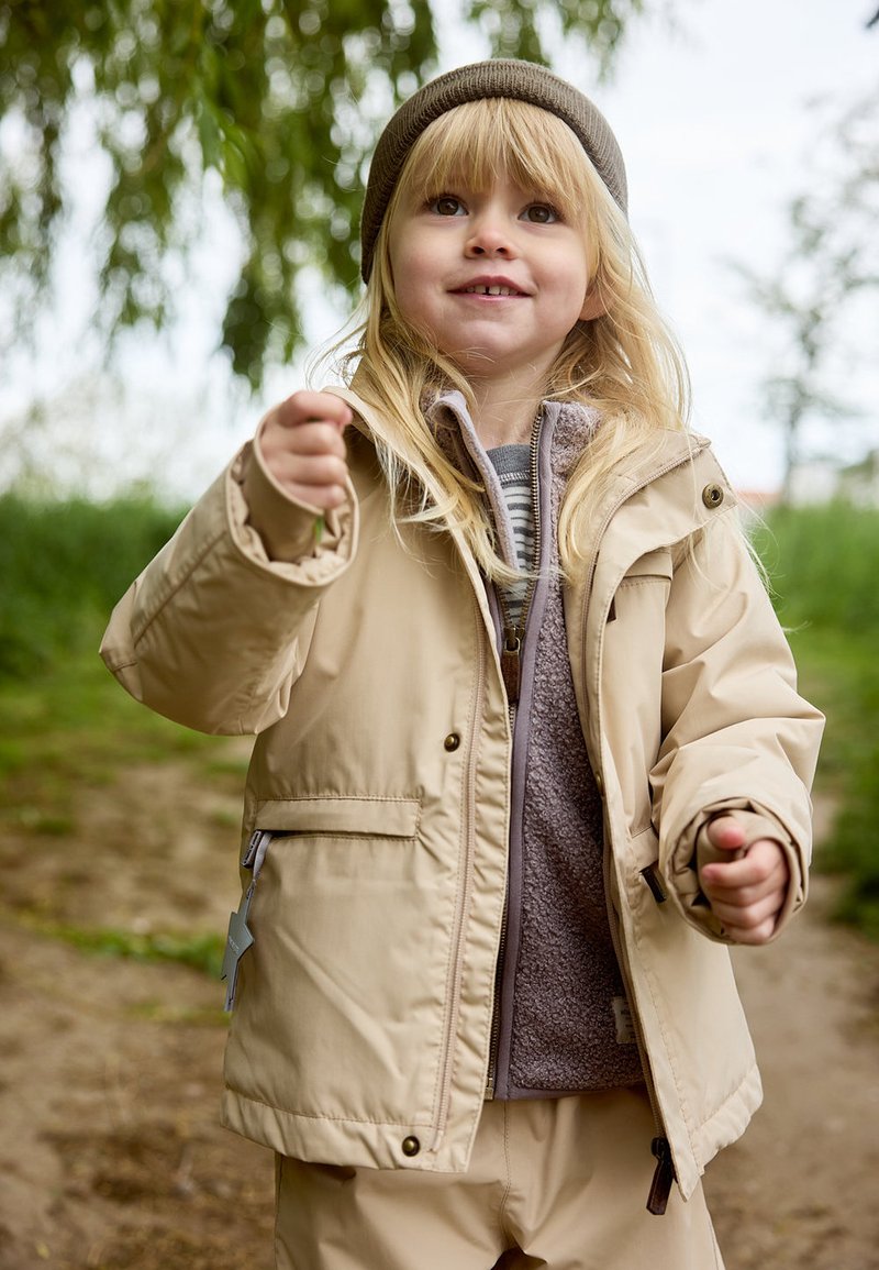 Giacca impermeabile beige con cappuccio, chiusura con zip e due tasche laterali, indossata sopra un maglione di pile grigio, indossata da un bambino all'aperto.