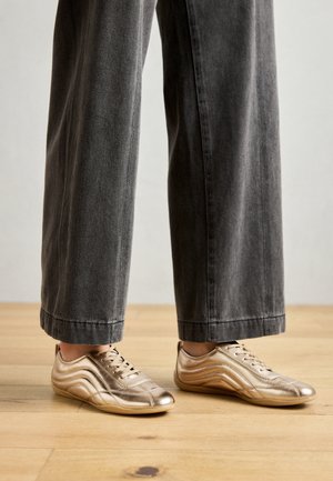 Fødder iført metalliske gyldne sneakers kombineret med vidtbenede mørkegrå bukser stående på et trægulv.