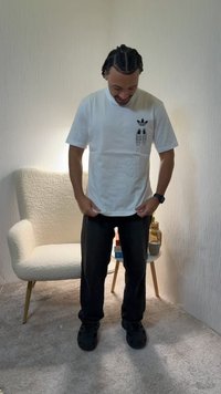 Mann med flettehår som har på seg en hvit Adidas-t-skjorte, svarte bukser og svarte sko, står i et teppebelagt rom med en lenestol og et lite sidebord.