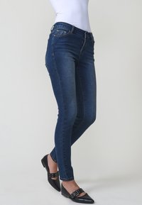 Femme portant un jean slim bleu foncé associé à des ballerines noires à bout pointu avec des brides à boucle, debout devant un fond clair uni.