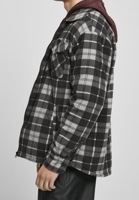 Veste en flanelle à carreaux noire et grise, avec une fermeture éclair sur le devant, des poignets boutonnés et une capuche bordeaux contrastante. Texture douce avec deux poches poitrine.