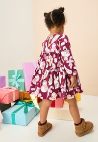 Lila klänning med festliga djurmönster, långa ärmar och en utsvängd kjol. Bruna stövlar och färgglada presentaskar i bakgrunden.