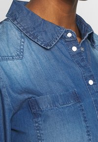 Denim-paita, jossa on vaaleansininen sävy, klassinen kaulus, nappikiinnitys valkoisilla napeilla ja rintatasku. Tasainen pinta ja rento muotoilu.