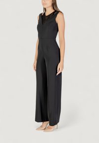 Mono negro con detalles de encaje en el escote, diseño sin mangas, corte de pierna ancha y textura suave. Modelo llevando tacones nude.
