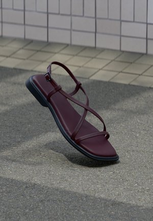 Σανδάλι από σκούρο κόκκινο δέρμα με λεπτά λουράκια που διασταυρώνονται και μαύρη σόλα, ακουμπισμένο σε γκρι πεζοδρόμιο κοντά σε τοιχο με πλακάκια.