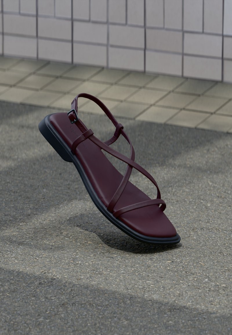 Sandale en cuir rouge foncé avec des fines lanières croisées et une semelle noire posée sur un trottoir gris près d’un mur carrelé.