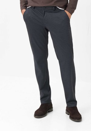 Homme portant un pantalon à carreaux gris foncé, un pull marron et des bottes en daim marron foncé, debout les mains dans les poches sur fond blanc.