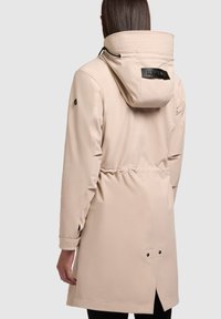 Parka imperméable beige avec capuche à design côtelé, cordon de serrage, taille élastique et boutons-pression. Texture lisse.