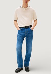 Polo beige con dettagli neri, jeans in denim blu, cintura nera intrecciata e mocassini neri, in piedi con le mani nelle tasche.