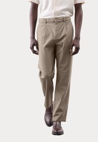 Pantalones beige de corte holgado con pliegues en la parte frontal, con cierre de botón y bolsillos laterales, combinados con zapatos marrón oscuro.