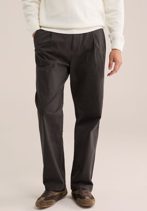 Mann trägt dunkelgraue Hose mit geradem Bein, braune Wildleder-Schnürschuhe und einen cremefarbenen Strickpullover mit einer Hand in der Tasche.