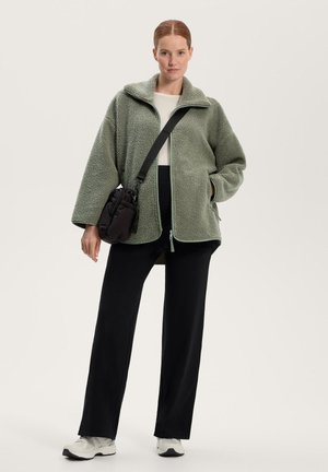 Flis jakna svetlo zelene barve, zasnova oversized z zadrgo in stranskimi žepi. Kombinirana s črnimi hlačami in belimi supergami.
