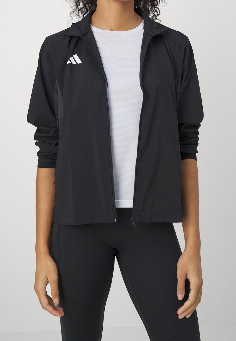 Personne portant une veste noire zippée avec un logo blanc sur une chemise blanche et un pantalon noir, debout devant un fond clair uni.