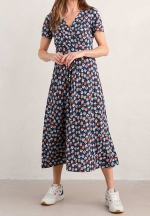 Femme portant une robe midi bleu marine à motif floral avec manches courtes et des baskets blanches, debout devant un fond beige uni.