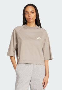 Svetle hnědý zkrácený tričko s krátkým rukávem a žebrovaným límcem. Na hrudi se nachází bílé logo Adidas. Měkká, hladká látka.