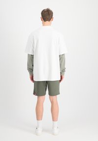 T-shirt oversize blanche superposée sur une chemise à manches longues couleur olive, associée à un short olive. Le mannequin se tient de dos face à l'objectif.
