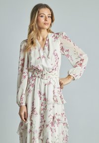 Lungo abito floreale in crema chiaro, con motivi di fiori rosa, volant e vita cinturata. Le maniche lunghe sono leggermente gonfie.