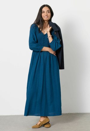 Woman wearing a long blue dress and brown shoes, holding a dark coat over one shoulder, standing against a plain white wall.